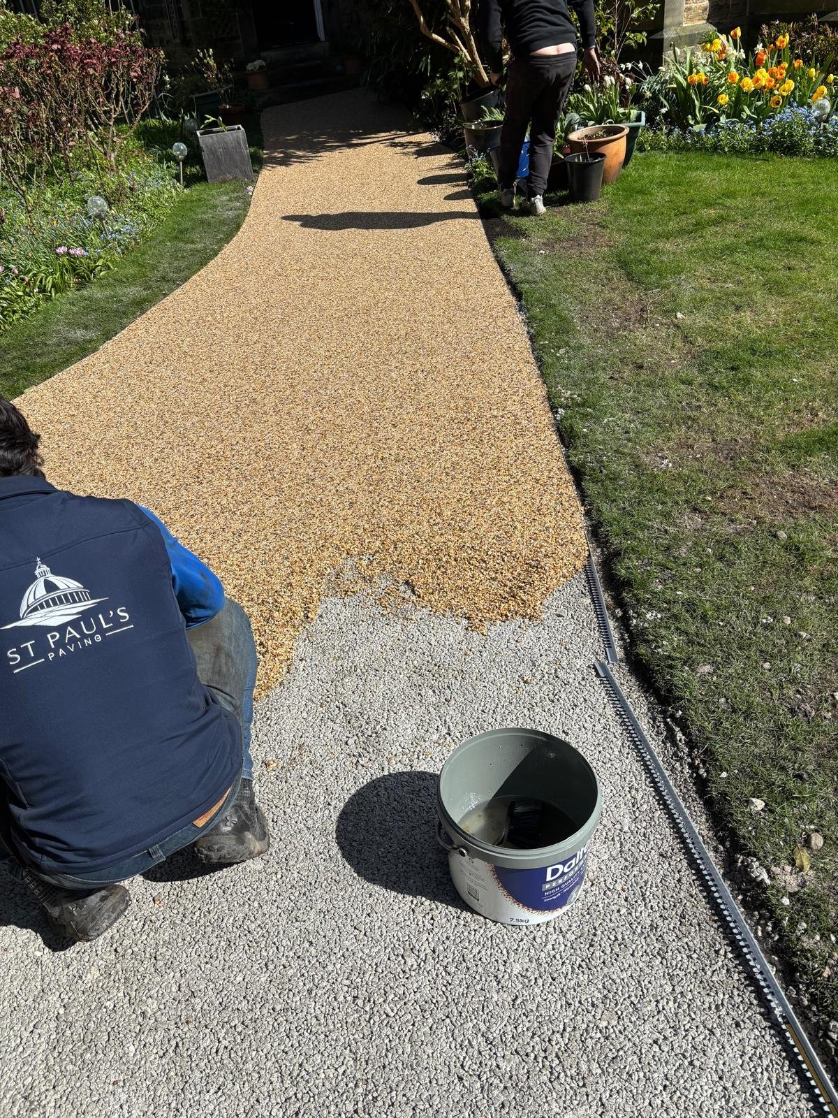 Worker installing tan and gray pebbled pathway in garden with flowering plants and green lawn visible